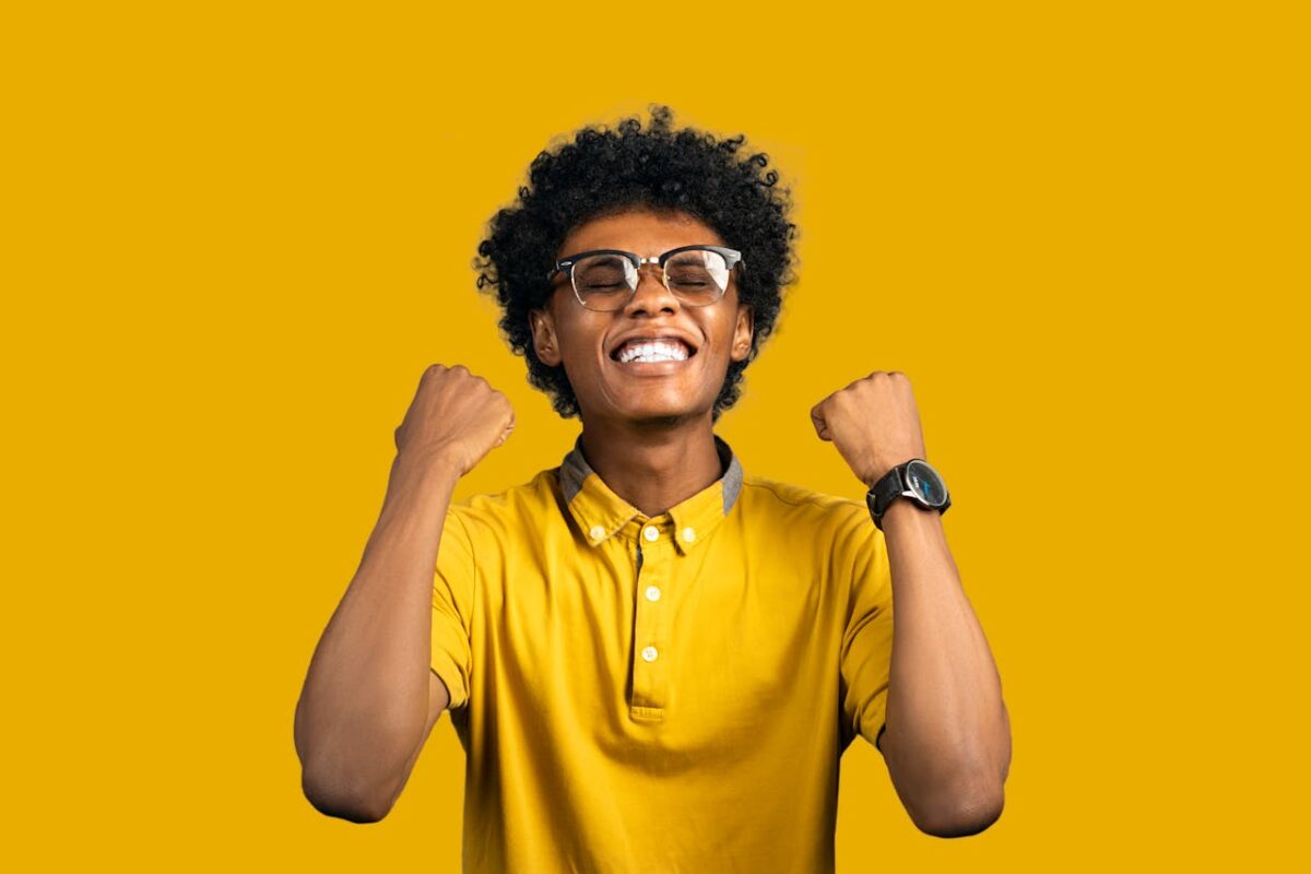 Happy young black male in bright t shirt and eyeglasses with Afro hairstyle standing with closed eyes while demonstrating achievement gesture on yellow background
